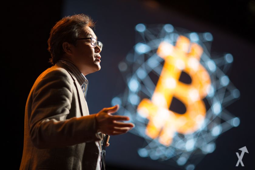 Tom Lee falando sobre Bitcoin em uma conferência, logo do Bitcoin ao fundo.